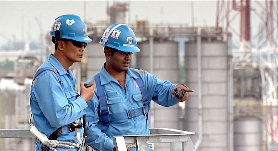 Two workers in blue overalls and hard hats at a refinery
